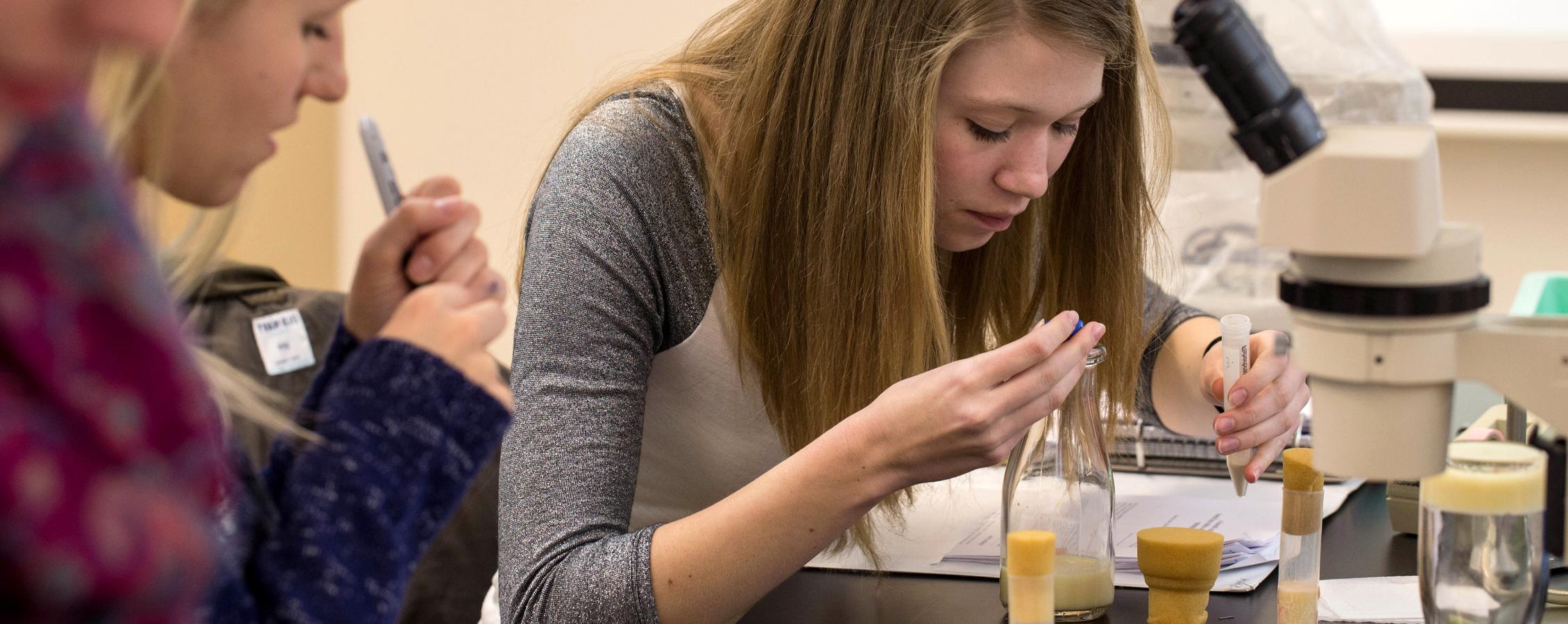 A student inspects something in a vial next to a microscope.