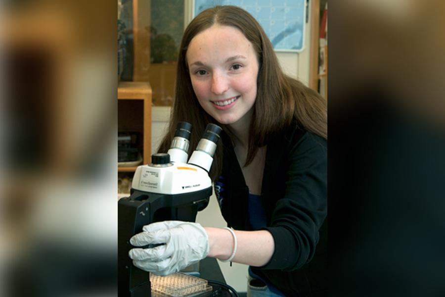 A woman looks up from a microscope and smiles at the camera.