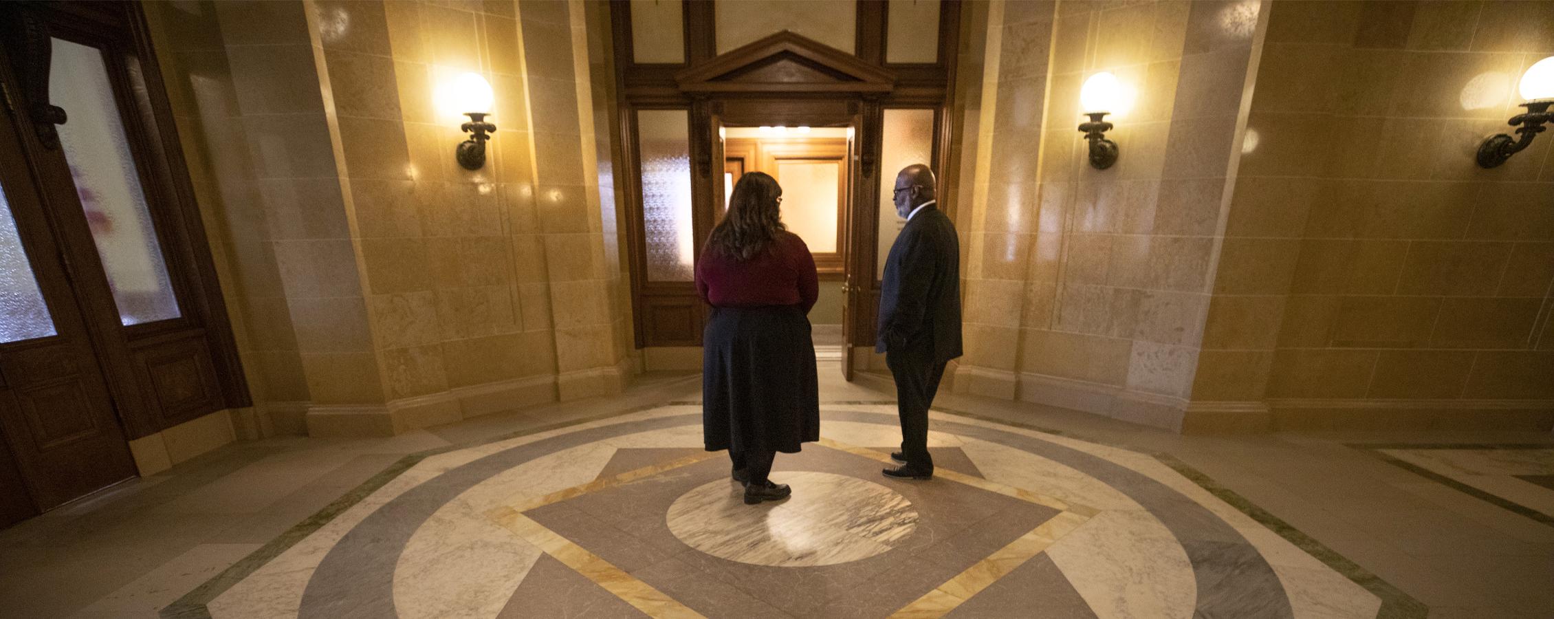 Two people talk in a room with dark woodwork and and American flag.