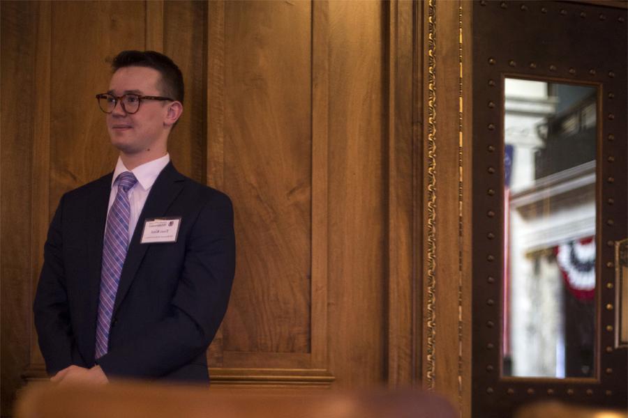 A student wearing a nametag stands against a beveled wood paneled wall.