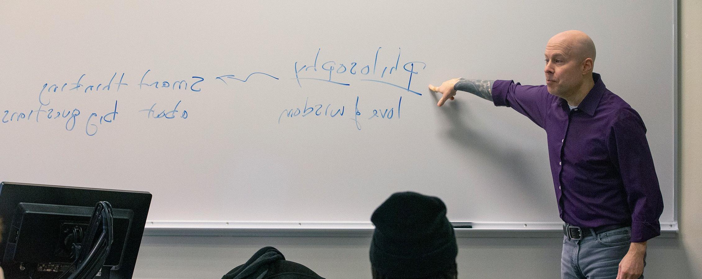 Chris Minor, professor of philosophy, pointing to the word Philosophy on a whiteboard.