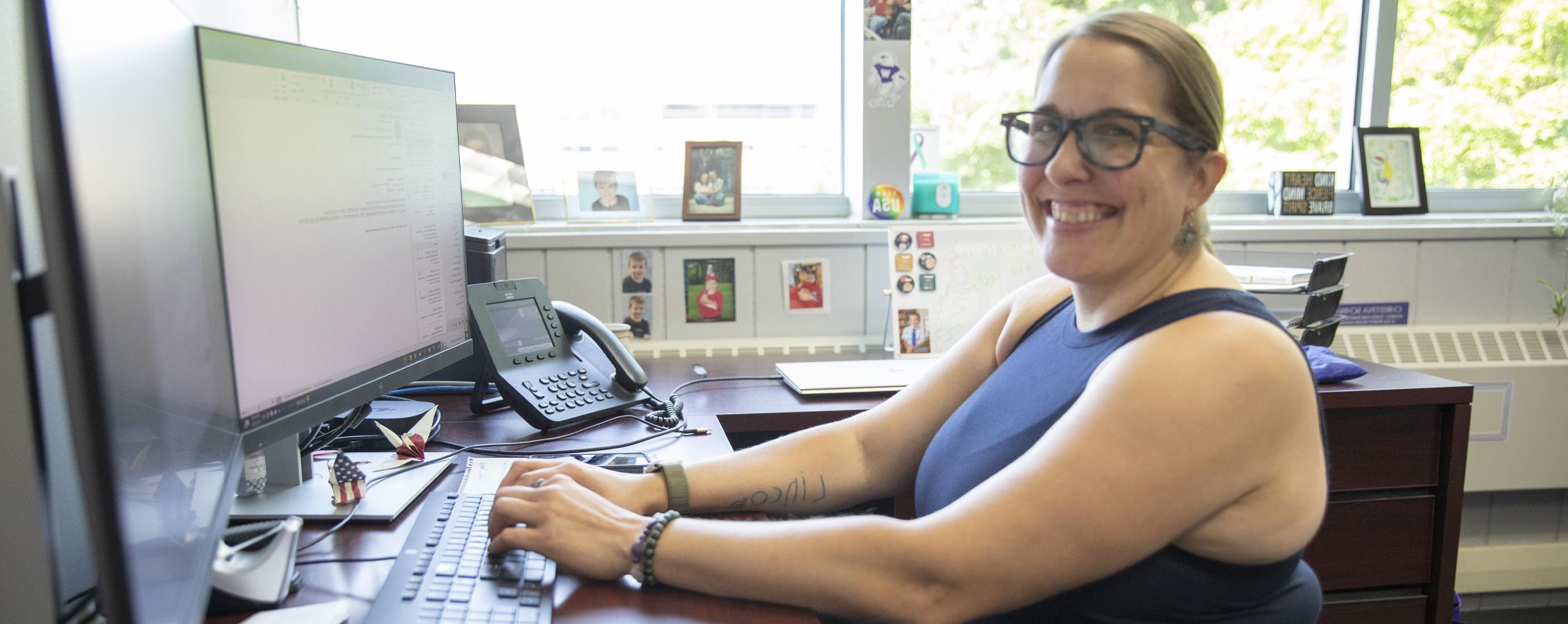 A person sits at desk with their hands on the keyboard and smiles at the camera.