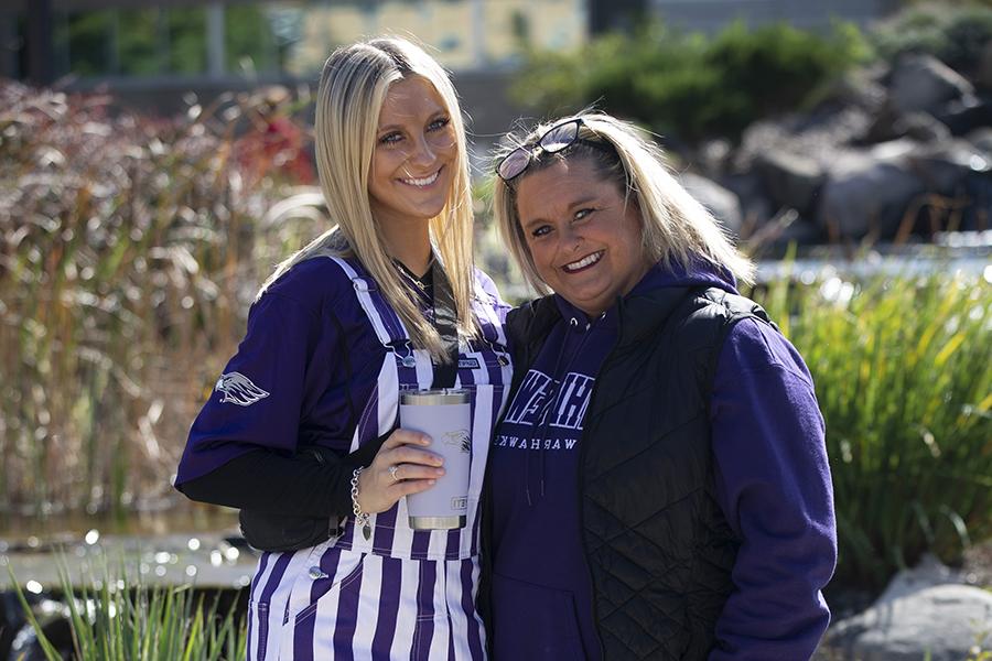 Two women outside at a tailgate at a football game, one wearing a purple sweatshirt and one in purple and white overalls.