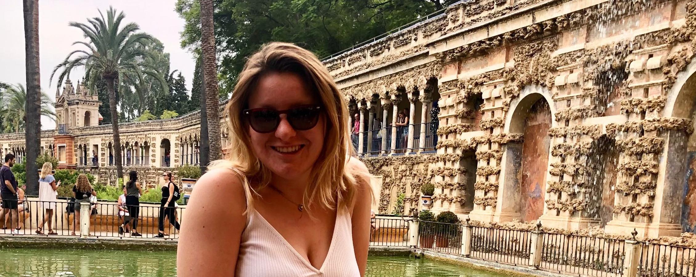 A young adult who is standing amidst a mediterranean style building smiles at the camera.