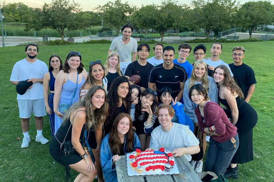 A group of international students celebrate with a red, white, and blue cake in a park.