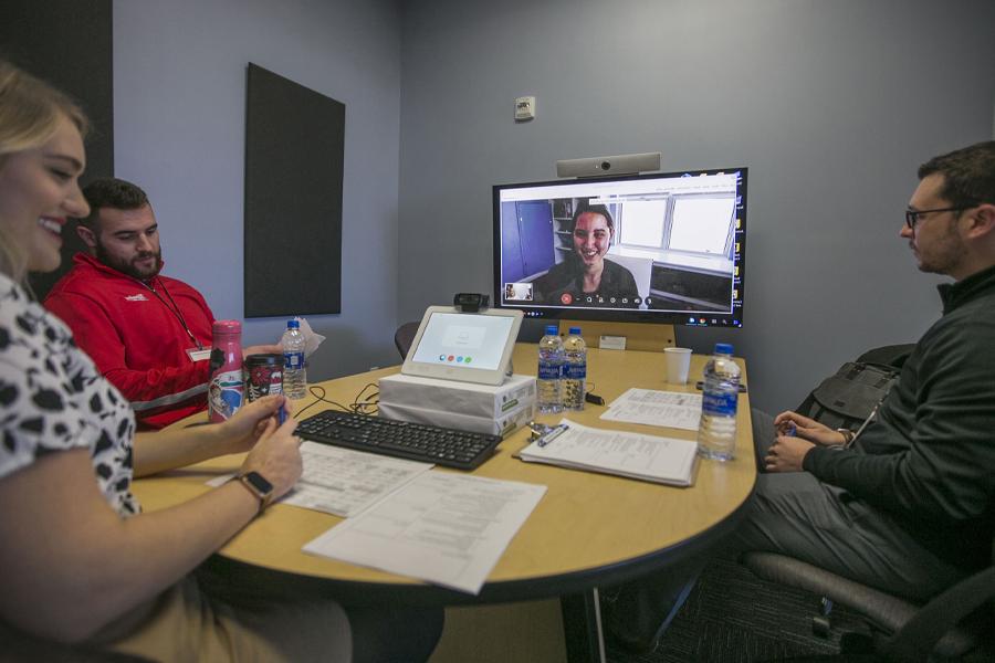 Three people sit around a table with a monitor during a video conference.