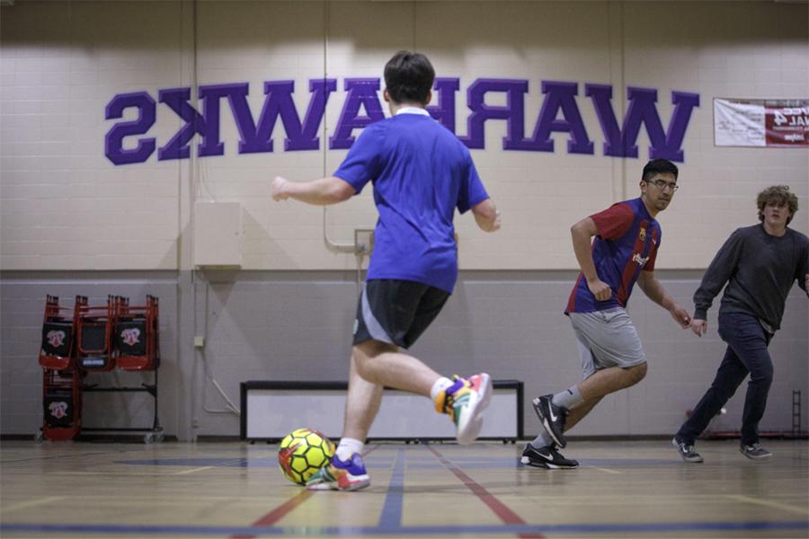 A student dribbles a soccer ball in a gym with the words Warhawks on the wall.